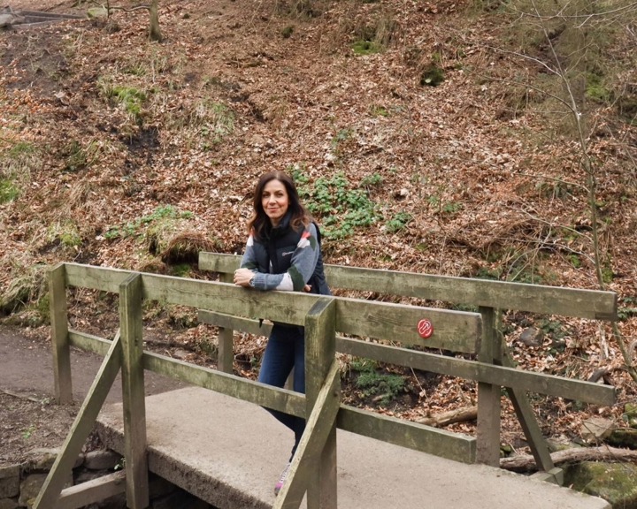 A person stands on a small wooden bridge in a forested area, leaning on the railing and looking toward the camera. The bridge spans a narrow stream, with fallen leaves and sparse vegetation covering the ground. A hillside with trees and underbrush rises in the background.