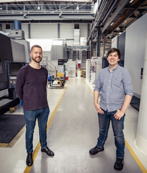 Two people standing in a spacious industrial workshop with large machinery on both sides. The environment features high ceilings, overhead beams, and bright lighting. The floor has marked yellow lines for pathways, and various equipment and workstations are visible in the background.