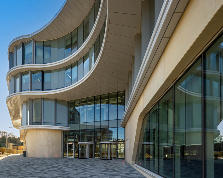Modern building with a striking curved glass façade and multiple levels featuring sweeping architectural lines. The entrance has large glass doors beneath an overhang, and the exterior includes reflective windows and light-coloured panels. The paved area in front of the building leads to an open outdoor space under a clear blue sky.