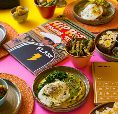A colorful table setting featuring several plates and bowls of assorted dishes, including eggs on greens, fried vegetables, and small sides with sauces and blueberries. Two illustrated hip-hop themed books are placed among the food, and the table is covered with bright pink and yellow surfaces accented by orange placemats.
