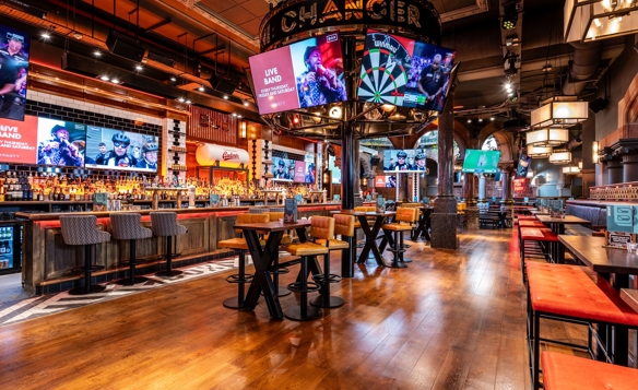 A spacious, modern bar interior with wooden floors, high tables, and cushioned stools. Multiple large screens display sports and live band promotions. The central feature is a circular structure above the bar with a large dartboard graphic. Warm lighting and industrial-style décor create an energetic atmosphere.