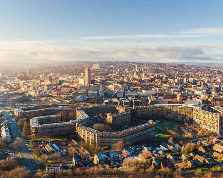 Aerial view of Sheffield cityscape under clear skies, showing a mix of modern and historic buildings. Prominent in the foreground is the curved Park Hill housing complex with its distinctive architecture, surrounded by roads and greenery. The city centre stretches into the distance with tall buildings, including the central tower, and rolling hills visible on the horizon.