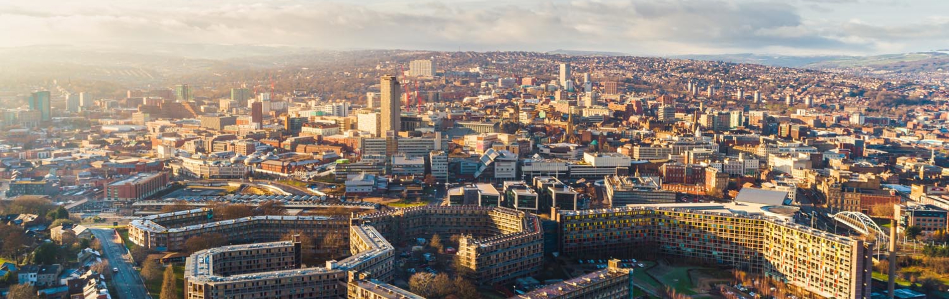 Aerial view of Sheffield cityscape under clear skies, showing a mix of modern and historic buildings. Prominent in the foreground is the curved Park Hill housing complex with its distinctive architecture, surrounded by roads and greenery. The city centre stretches into the distance with tall buildings, including the central tower, and rolling hills visible on the horizon.
