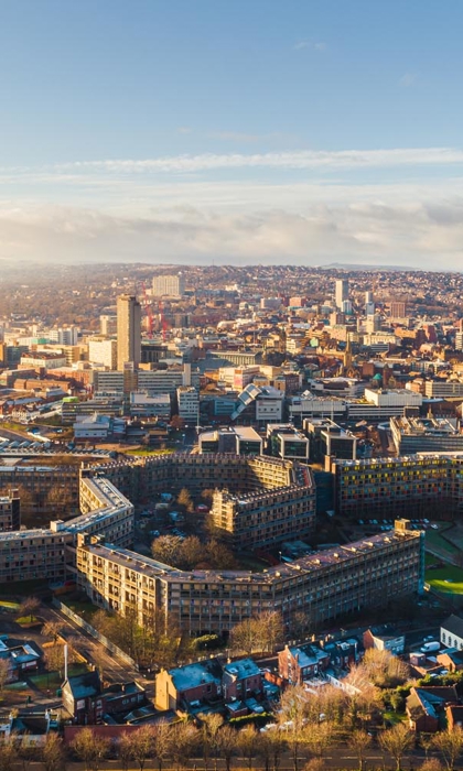 Aerial view of Sheffield cityscape under clear skies, showing a mix of modern and historic buildings. Prominent in the foreground is the curved Park Hill housing complex with its distinctive architecture, surrounded by roads and greenery. The city centre stretches into the distance with tall buildings, including the central tower, and rolling hills visible on the horizon.