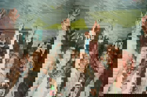 A lively indoor party scene with several people raising their arms in the air, holding drinks and cups. The background features colorful decorations, including draped fabric on the ceiling and a large banner with partially visible text. There is also an inflatable figure positioned on the left side of the image. The setting appears festive and crowded.