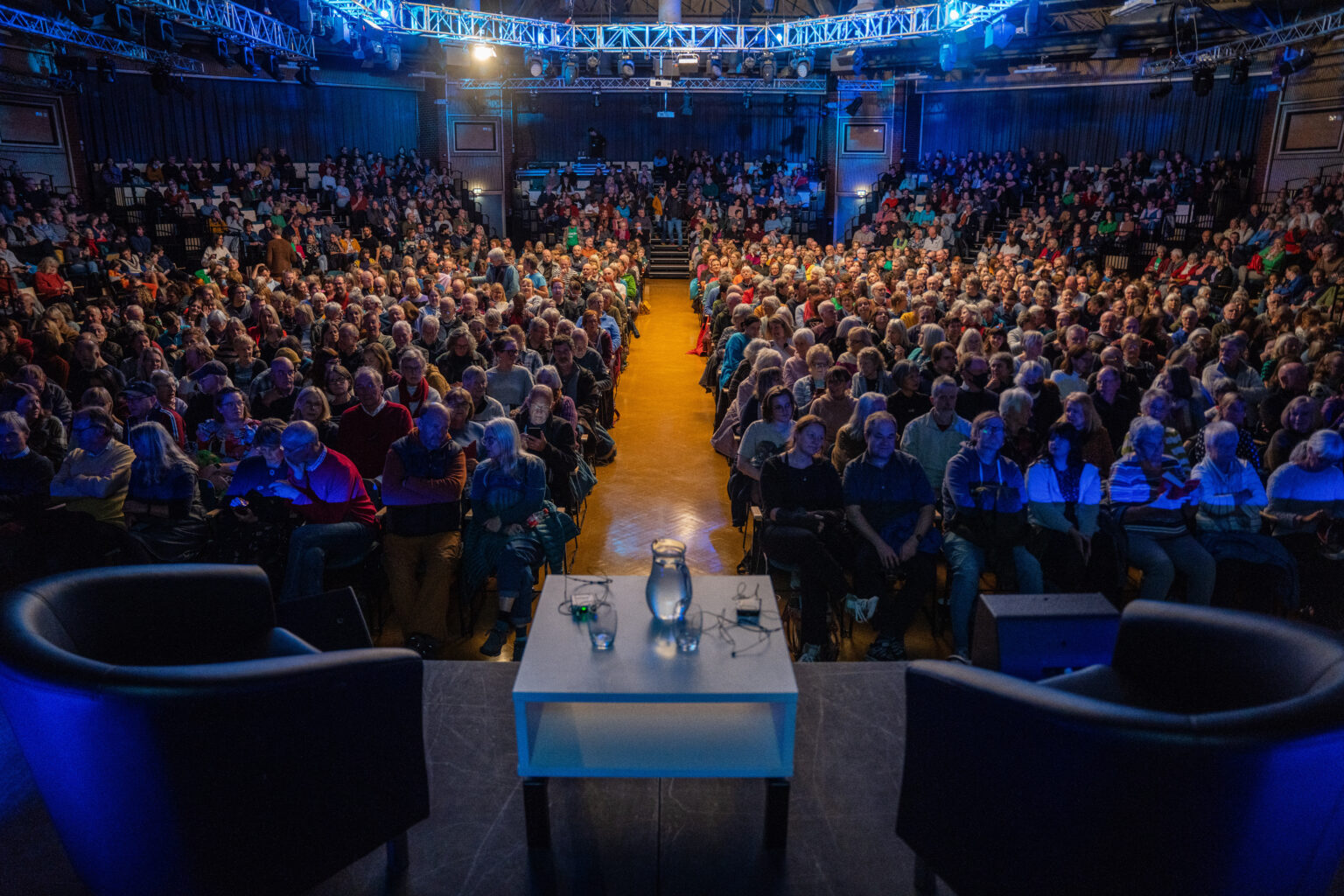 A large audience is sat waiting for an event to begin. On the stage are two armchairs on either side of a low table that has two glasses and a pitcher of water sat on it.