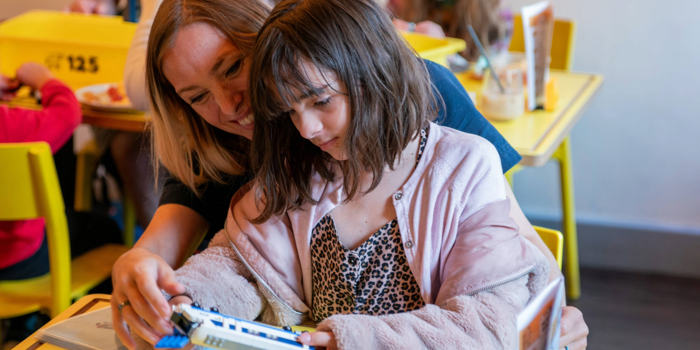 A person and a child are sat at a wooden table in a cafe, building a Lego© train together.