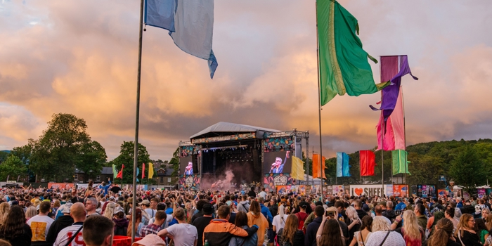 Large outdoor music festival with a crowd gathered in front of a main stage during sunset. The stage features large screens showing performers, and colorful flags in blue, green, purple, orange, and red are positioned throughout the field. A sign reading ‘I ❤️ SHEFF’ is visible near the stage area, with trees and hills in the background under a dramatic cloudy sky.
