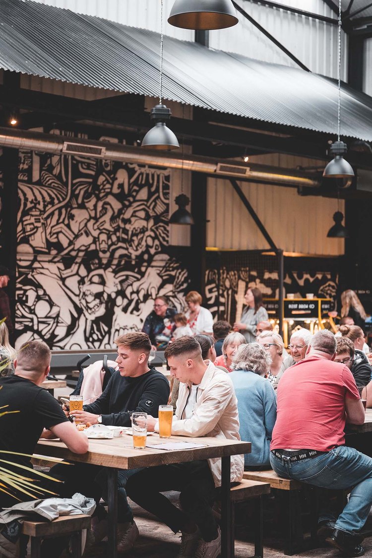 Crowds of people sat on benches drinking beer indoors, with a black and white graffiti wall in the background