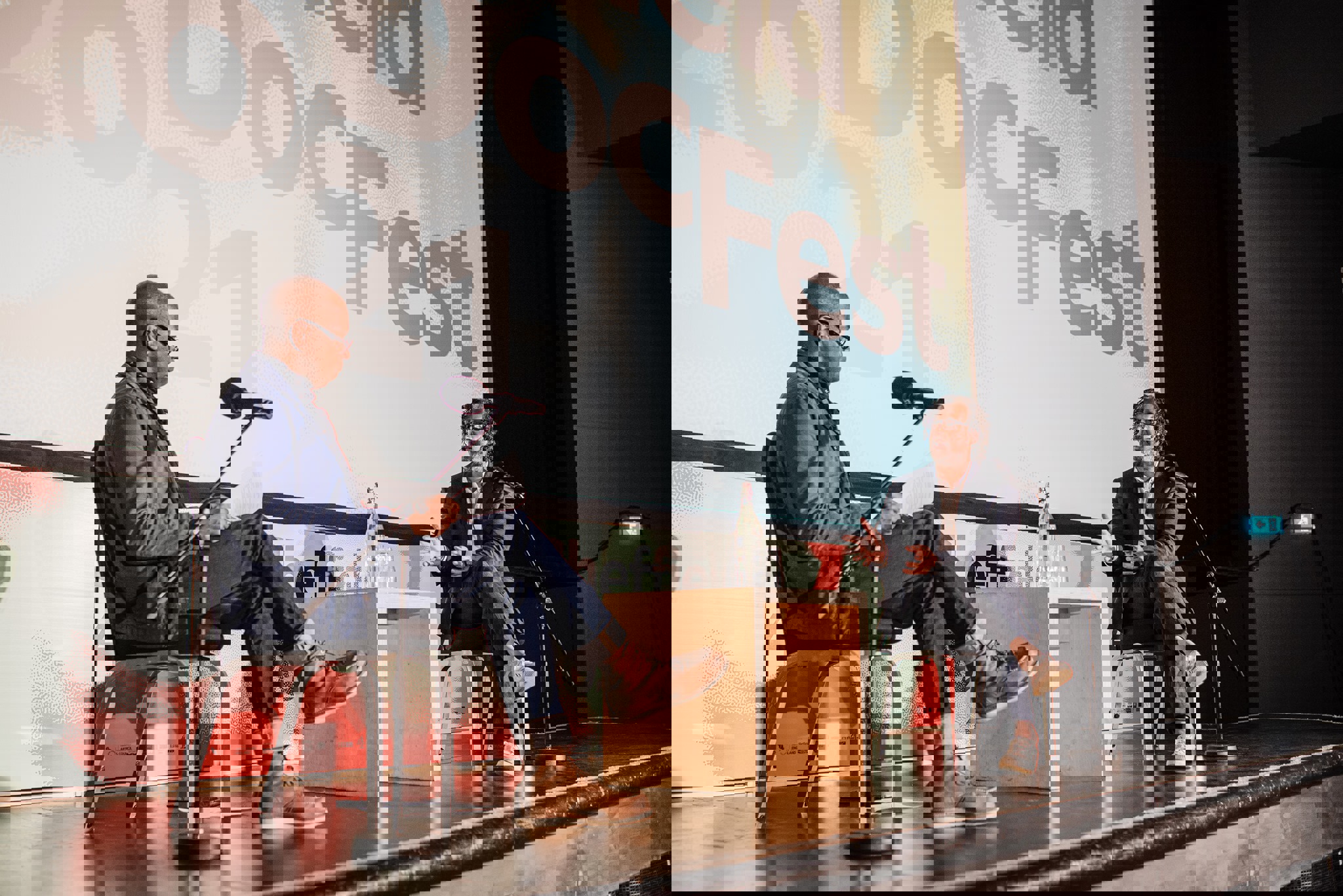 Two people seated on stage during a discussion at Sheffield DocFest, with microphones on stands and a small wooden table between them. A large screen behind displays the event name and year.