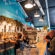 Two people are behind a bar, pulling pints of beer, as two customers wait on the other side.