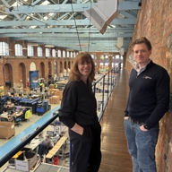 Two people standing on an upper-level walkway inside a large industrial building with exposed brick walls and blue-painted steel beams. The walkway overlooks a spacious factory floor filled with workstations, machinery, and shelving units. The area below is organized with tables, equipment, and workers in high-visibility clothing. Large arched windows line the brick walls, allowing natural light into the space.