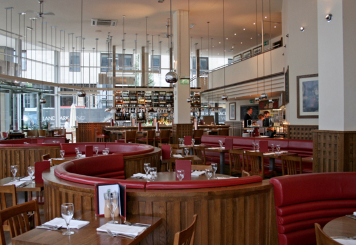 The interior at Piccolino. Bright, modern restaurant interior with large windows, high ceilings, and hanging pendant lights. The space features curved wooden booths with red leather seating and neatly set tables with glassware and menus. A long bar at the back is stocked with bottles, and the overall design combines warm wood tones with contemporary elements.