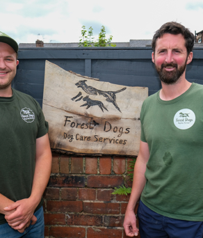 Two men are stood next to a fence that has a sign on itthat reads 'Forrest Dogs'.