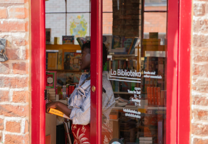 View through a red-framed glass door into a bookshop called La Biblioteka. Inside, shelves are filled with colourful books and magazines. A person is holding a yellow book titled “CRAFTLAND” while browsing. The shop’s name and opening hours are printed on the glass, and the interior appears bright and inviting with a variety of reading materials on display.