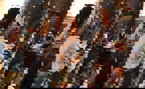 A cozy indoor setting with several people gathered on a sofa and nearby chairs, holding paper cups and food trays. Warm string lights hang overhead, and the background shows exposed brick walls and soft ambient lighting, creating a relaxed social atmosphere.