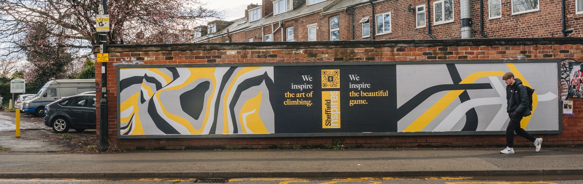 A street mural painted on a brick wall featuring abstract patterns in black, white, gray, and yellow. The mural includes two text sections: “We inspire the art of climbing” and “We inspire the beautiful game.” A person wearing a hooded jacket and carrying a backpack walks past the mural on the sidewalk. Behind the wall are rows of red-brick houses with white-framed windows and sloped roofs. The street in front has faded yellow road markings and appears wet, suggesting recent rain.