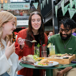 Three people seated at a round green table in an indoor food hall, sharing a meal. The table holds plates of tacos, salad, bread, and a wooden tray with assorted dishes. There are also tall glasses of colourful drinks garnished with fruit and herbs. In the background, green triangular banners hang overhead, and signs for “Oktoberfest” and “Baity” are visible, creating a lively, casual dining atmosphere.