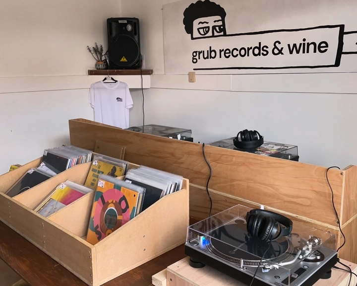 Interior of a small record shop with wooden bins filled with vinyl records, a turntable with headphones on the counter, and a wall sign reading ‘grub records & wine’ above a hanging white T-shirt.