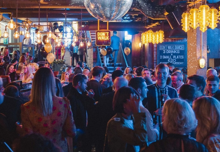 Manahatta filled with people. Crowded bar scene with a large disco ball hanging from the ceiling, surrounded by warm pendant lights and gold balloons. People are gathered closely, chatting and holding drinks. A neon-style sign reading “DON’T WALK” hangs above the crowd, and a chalkboard menu on the right advertises drink and food specials. The atmosphere is lively with dim, colourful lighting and industrial-style decor.