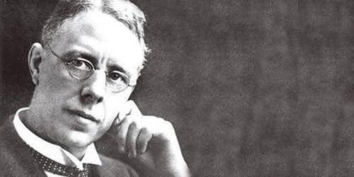 Black-and-white photograph of a person wearing a formal suit with a collared shirt and tie, seated and resting one arm on a surface while the other hand is positioned near the chin. The background is plain and dark, creating a classic portrait style.