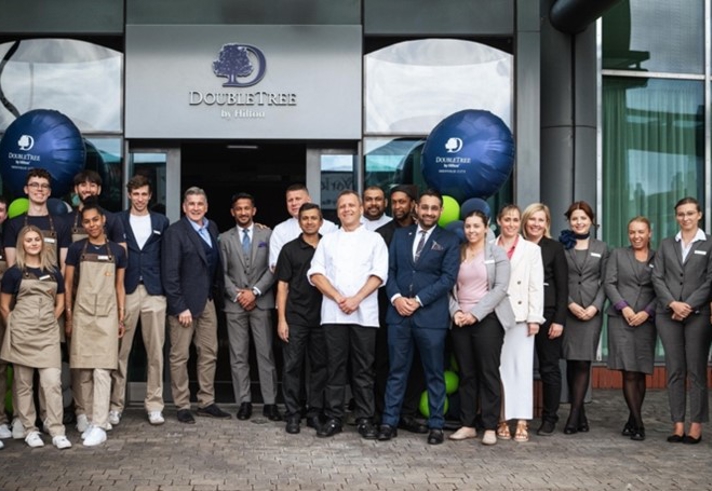Group of people standing in front of the entrance to a DoubleTree by Hilton hotel, posing for a photo. The group includes individuals wearing business attire and others in casual uniforms, with two large branded balloons on either side of the doorway.