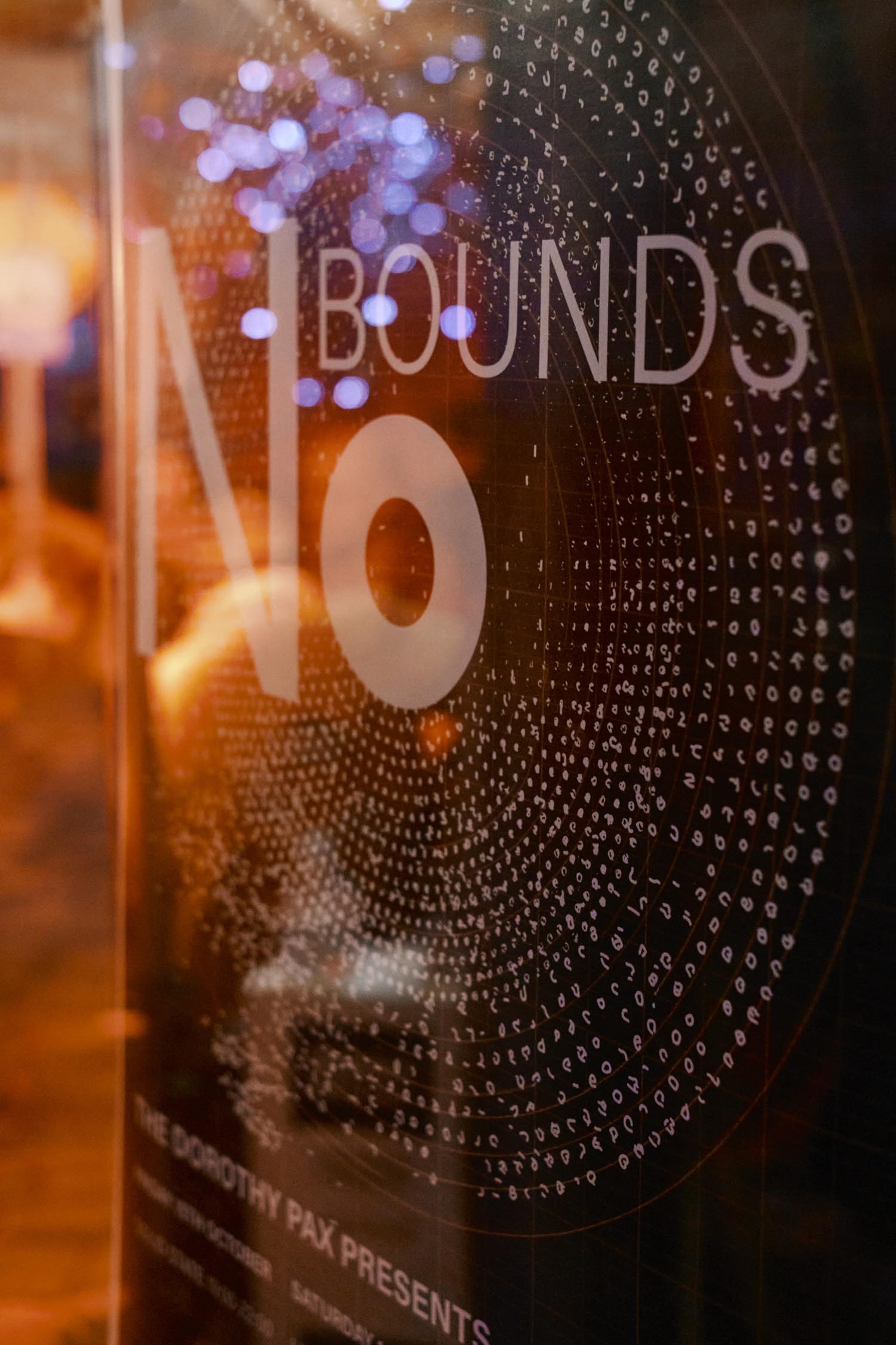 A close-up of a glass panel with the words “NO BOUNDS” displayed in large white letters, accompanied by a circular pattern of small characters radiating outward. The design appears artistic and modern, with warm orange reflections and purple bokeh lights in the background. The bottom of the panel includes smaller text mentioning “The Dorothy Pax Presents,” suggesting an event or promotional poster.