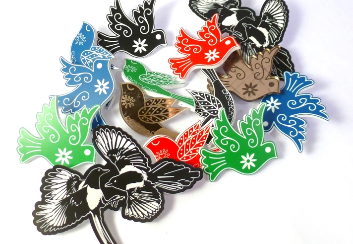 Collection of colorful bird-shaped brooches in red, green, blue, black, and metallic tones with intricate floral and leaf patterns, arranged on a white background.