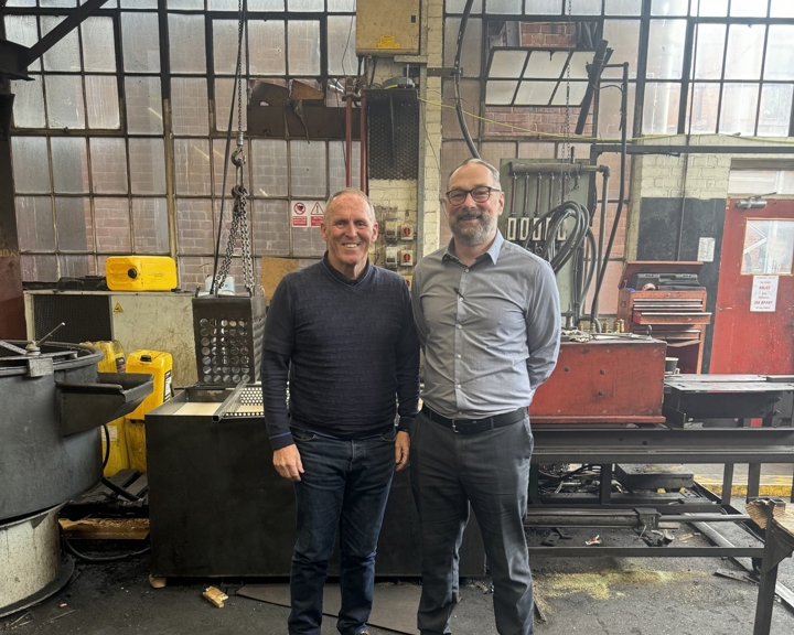 Two people standing inside an industrial workshop with large multi-pane windows in the background. The space contains heavy machinery, metal workstations, chains, and equipment, including a red tool cabinet and yellow safety blocks. The floor is dark and worn, and the overall setting suggests a manufacturing or engineering environment.