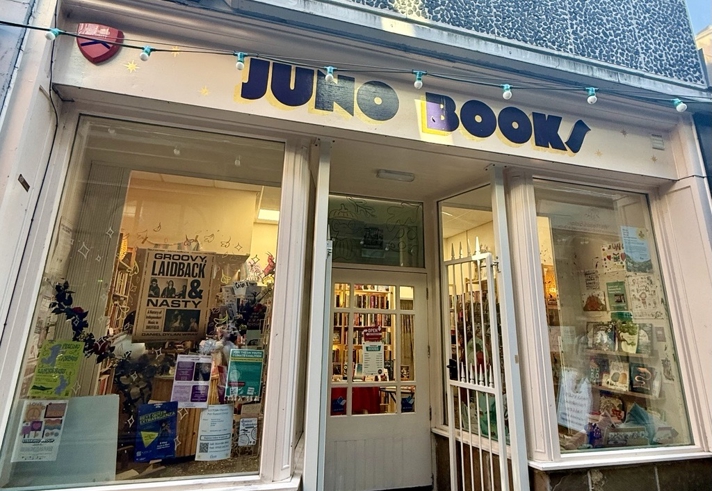 The exterior of an independent bookshop called “Juno Books,” featuring large front windows filled with posters and displays. The shopfront is cream-coloured with a central doorway leading inside, and a string of turquoise fairy lights hangs above the entrance.