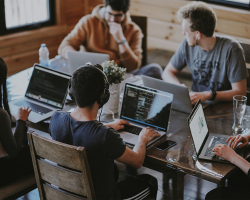 Five remote workers in casual dress sit round a large table, working on laptop computers.