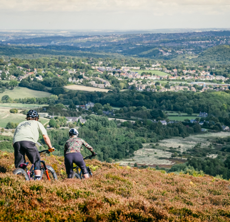 Two people are mountain biking along a dirt track, flanked by heather, on a hillside above Sheffield.