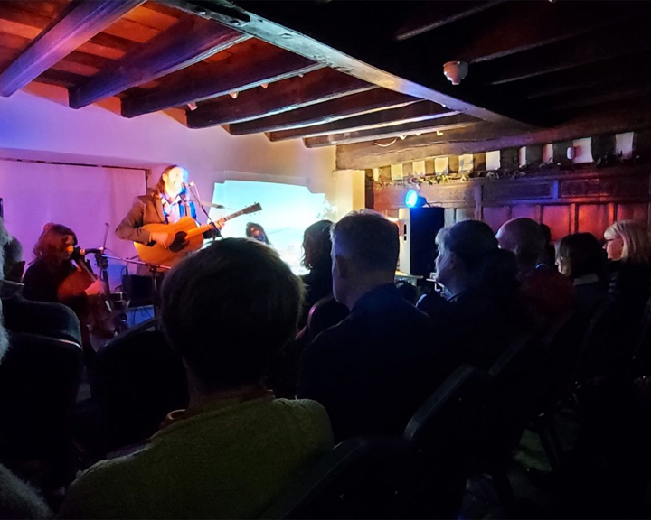 Indoor live music performance in a rustic venue with exposed wooden beams and warm lighting. A musician is playing an acoustic guitar and singing on a small stage, illuminated by blue and purple lights. A cello player is seated nearby. The audience is seated in rows of chairs, watching the performance. The background includes wooden panelling and a projector screen displaying a faint image.