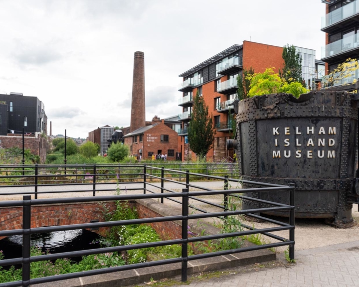 Outdoor view of Kelham Island Museum in Sheffield. A large industrial metal container with “Kelham Island Museum” written on it stands in the foreground near railings and a water channel. Surrounding the area are modern apartment buildings, a tall brick chimney, and greenery under a cloudy sky.