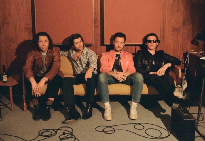 Four people sitting on a mustard-yellow sofa in a wood-paneled room with acoustic panels on the wall. They are dressed in casual clothing, including leather jackets, striped shirts, and a bright coral jacket paired with jeans and white sneakers. The setting includes musical equipment such as an amplifier and cables on the floor, along with a small side table holding a bottle. The overall atmosphere suggests a relaxed studio or rehearsal space.