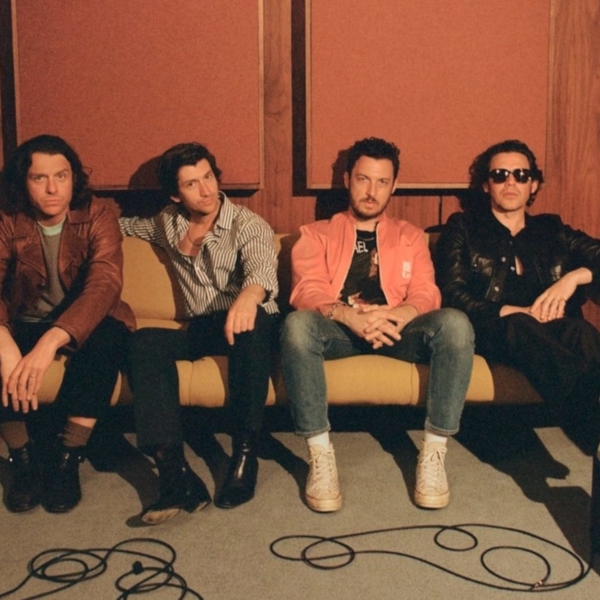 Four people sitting on a mustard-yellow sofa in a wood-paneled room with acoustic panels on the wall. They are dressed in casual clothing, including leather jackets, striped shirts, and a bright coral jacket paired with jeans and white sneakers. The setting includes musical equipment such as an amplifier and cables on the floor, along with a small side table holding a bottle. The overall atmosphere suggests a relaxed studio or rehearsal space.