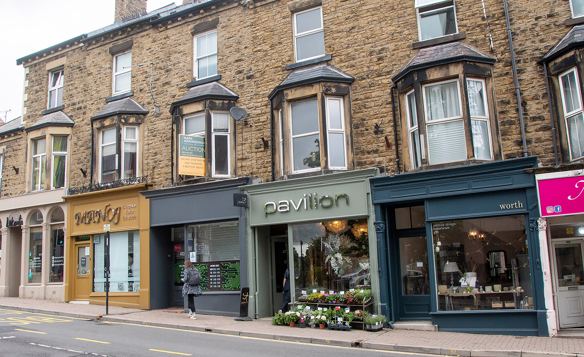 Row of stone-built Victorian-style buildings with ground-floor shops along a street. Visible storefronts include “Valley Insurance Services” with a mustard-yellow facade, “Pavilion” with green signage and flower display outside, and “Worth” with a dark blue front showcasing homeware items. A person stands near the insurance shop, and a “For Auction” sign is displayed in an upper window.