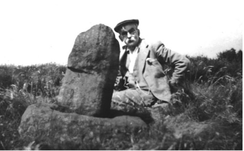 Black-and-white historical photograph showing a person seated outdoors on grassy terrain beside a large upright stone with a smaller stone at its base. The scene suggests a rural or archaeological setting, with natural vegetation in the background under bright daylight.