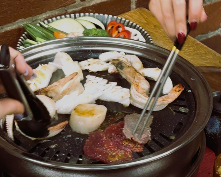 Close-up of a tabletop grill with various seafood and meat pieces cooking, including shrimp, scallops, and beef slices. Two hands are using tongs and chopsticks to turn the food. In the background, a plate of fresh vegetables such as zucchini, bell peppers, and green onions is visible.