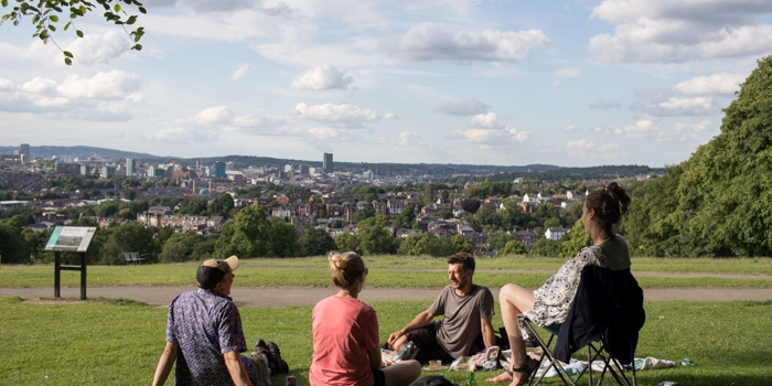 Four people are sat on the grass at Meersbrook Park in the sun. In the distance you can see Sheffield city centre.