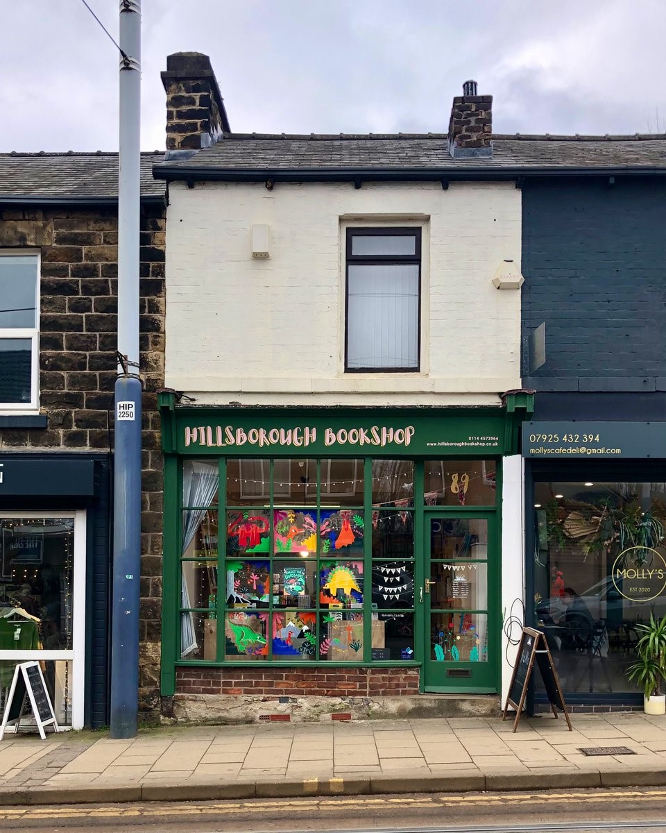Front-facing street view of Hillsborough Bookshop situated between neighbouring buildings. The shop has a green façade with a large display window filled with brightly coloured artwork, and the sign ‘HILLSBOROUGH BOOKSHOP’ is positioned above the window and door.