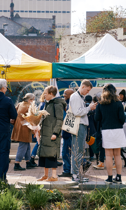 A lively outdoor market scene with people gathered around stalls covered by yellow and green canopies. Various individuals are browsing items, chatting, and holding bags or flowers. Display boards and tables with informational materials are set up under the tents. The background shows older brick buildings and a tall modern structure, while the foreground features plants and a paved area.