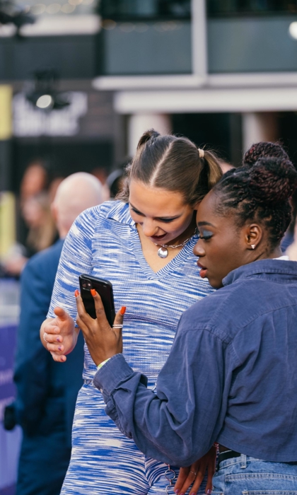 Two people standing close together outdoors, one holding a smartphone and showing the screen to the other. The person on the left is wearing a blue patterned outfit with a heart-shaped necklace, while the person on the right is dressed in a dark denim shirt. The background shows a blurred crowd and building structures.