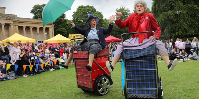 Two performers dressed in vintage-style clothing ride motorised shopping trolleys covered in tartan fabric during an outdoor event. One holds a bright green umbrella while both raise their arms in animated gestures. Behind them, a crowd watches near colourful market-style tents set up on a grassy area with trees and a large classical building in the background.