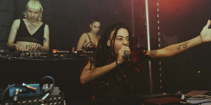 Three performers on stage at No Bounds festival. One is singing into a microphone while the other two are playing records on turntables and using a mixer.