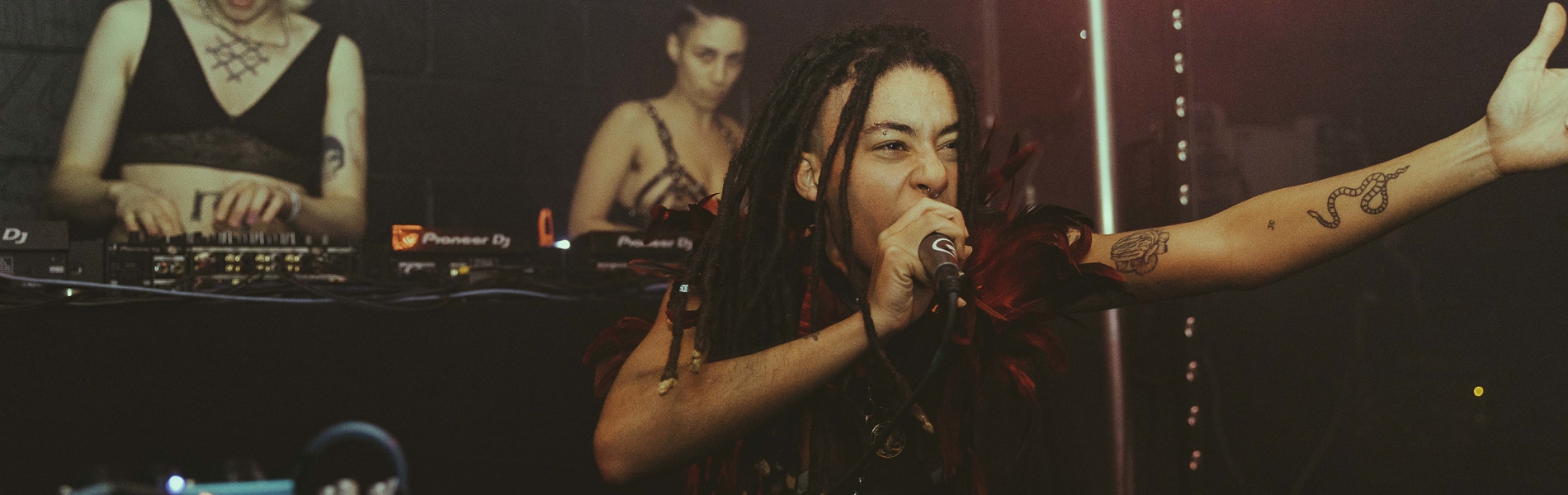 Three performers on stage at No Bounds festival. One is singing into a microphone while the other two are playing records on turntables and using a mixer.