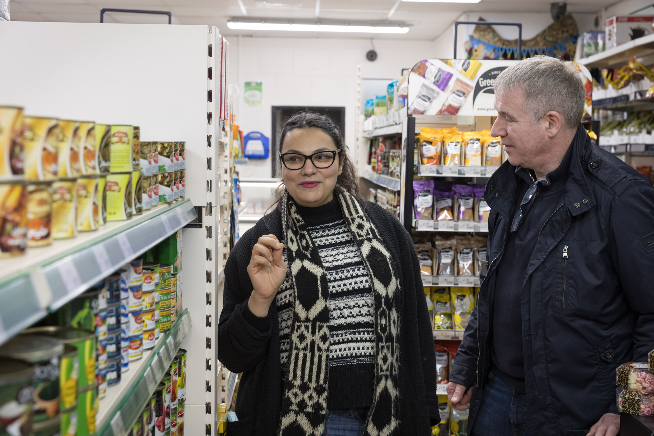 The inside of a shop  with a woman and a man standing in-between rows of shelves that are filled with food. 