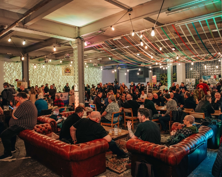 A large indoor festival space filled with people seated at tables and standing in groups. Colourful ribbons hang across the ceiling, and string lights illuminate the room. The atmosphere appears busy and social, with many attendees enjoying drinks.