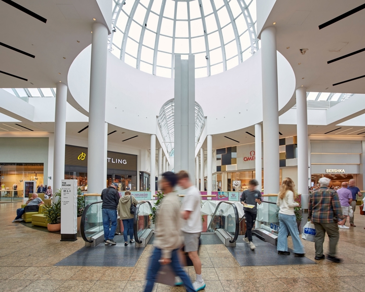 The interior of Meadowhall beneath a tall glass dome, showing escalators, white pillars, surrounding shops and shoppers moving through the space.