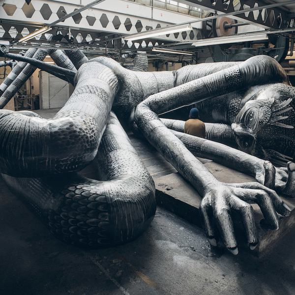 Large monochrome sculpture of a reclining humanoid figure with elongated limbs and intricate patterns, displayed inside an industrial-style warehouse with exposed beams and machinery.